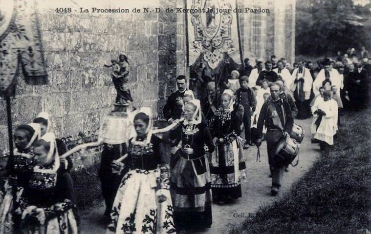 Period photograph of a Breton pardon procession in traditional dress passing a stone church wall